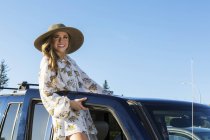 Улыбающаяся женщина в шляпе стоит рядом с открытой дверью автомобиля в дневное время — стоковое фото