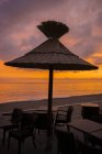 Ein Strohschirm über einem Tisch mit Stühlen am Strand bei Sonnenuntergang mit Blick auf das Mittelmeer; menton, cote d 'azur, Frankreich — Stockfoto