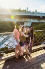 Quatro meninas felizes de pé na passarela de madeira contra esgrima de metal sobre a água e fazer selfie — Fotografia de Stock