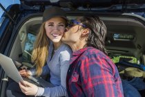 Усміхнена жінка в шапці з ноутбуком в руках сидить на машині, а чоловік цілує її — стокове фото