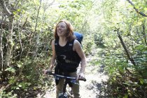 Una mujer se para en un camino en un bosque con su bicicleta y una mochila; Alaska, Estados Unidos de América - foto de stock