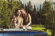 Two girls sitting on car top and hugging each other with trees on background — Stock Photo