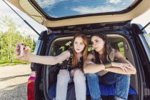 Две девушки сидят у машины и делают селфи на фоне деревьев — стоковое фото