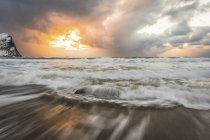 Flou de la marée se jetant sur le rivage sablonneux le long de la côte et un soleil doré culminant à travers les nuages orageux ; Nordland, Norvège — Photo de stock