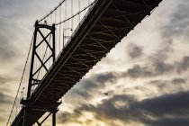 Vista de baixo ângulo de uma ponte ao pôr do sol com um avião voando na distância; Vancouver, Colúmbia Britânica, Canadá — Fotografia de Stock