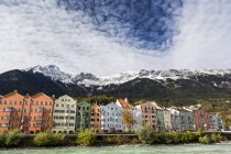 Edificios coloridos a lo largo de la orilla del río con picos de montaña cubiertos de nieve, nubes dramáticas y cielo azul; Innsbruck, Tirol, Austria - foto de stock