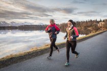 Duas jovens mulheres correndo em uma trilha na beira da água com montanhas à distância; Anchorage, Alasca, Estados Unidos da América — Fotografia de Stock