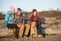 Ein junges paar und ein freund mit einem hund sitzen auf einem stück treibholz an einem strand mit blick auf das meer bei untergang; ankerplatz, alaska, vereinigte staaten von amerika — Stockfoto