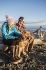 Um jovem casal com seu cão se senta em um pedaço de madeira à deriva em uma praia ao pôr do sol; Anchorage, Alaska, Estados Unidos da América — Fotografia de Stock