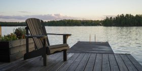 A wooden Adirondack chair sits on a dock on a tranquil lake at sunset; Lake of the Woods, Ontario, Canada — Stock Photo