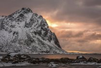 Rugged, snow-covered mountains with glowing pink clouds at sunset along the coastline of Norway; Nordland, Norway — Stock Photo