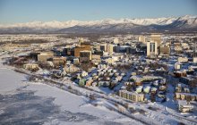 Aerial view of snow covering the sea ice on the frozen shores of downtown Anchorage, the Chugach Mountains in the distance beyond the office buildings and hotels, Cook Inlet in the foreground, South-central Alaska in winter; Anchorage, Alaska — Stock Photo