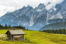 Grange en bois au sommet d'une prairie alpine avec une chaîne de montagnes accidentée en arrière-plan ; Sesto, Bolzano, Italie — Photo de stock