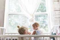 Two sisters playing together in crib by window — Stock Photo