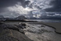 Un paisaje con montañas escarpadas y arena a lo largo de la costa bajo un cielo nublado; Nordland, Noruega - foto de stock