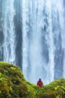 Homem sentado em uma rocha musgosa na base da cachoeira Skogafoss; Islândia — Fotografia de Stock