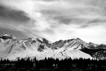 Black and white image of a snow covered mountain range under a cloudy sky with a forest in a valley; Mammoth Lakes, California, United States of America — Stock Photo