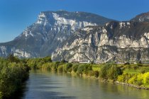 Tranquil river lined with trees in green foliage and rugged mountains in the background with blue sky; Trento, Trento, Italy — Stock Photo