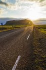 Солнце садится за холмы с асфальтированной дорогой, ведущей в закат, Исландия — стоковое фото