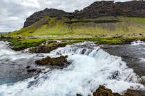 Largo ângulo do rio em cascata sobre as rochas em frente a uma montanha vulcânica e terras agrícolas, Islândia — Fotografia de Stock