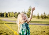 Cute young girl blowing a bubble and trying to catch it in a park — Stock Photo