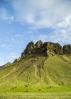 Прочный скалистый пик, который выглядит как памятник зеленый склон холма и голубое небо, общий вид на дорогу через Исландию, Исландия — стоковое фото
