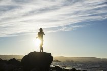A woman standing on a rock looking out along the coast at sunset, silhouetted and backlit by the sunlight; San Mateo, California, United States of America — Stock Photo