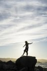 Una donna in piedi in equilibrio su un piede su una roccia con vista sulla costa al tramonto, sagomata e retroilluminata dalla luce del sole, San Mateo, California, Stati Uniti d'America — Foto stock