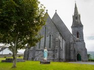 Iglesia de piedra con campanario y estatua de la mujer rezando en frente; Condado de Clare, Irlanda - foto de stock