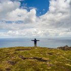 Femme debout avec les bras tendus et regardant l'océan et le littoral pendant une randonnée à Brandon Point, péninsule de Dingle ; Castlegregory, comté de Kerry, Irlande — Photo de stock
