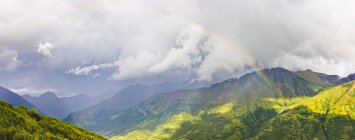 Arco iris que brilla a través de la luz atmosférica, exuberantes laderas verdes de las montañas en el fondo, Hatcher Pass, South-central Alaska; Palmer, Alaska, Estados Unidos de América - foto de stock