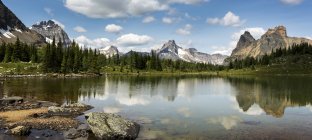 Vista panoramica della catena montuosa che si riflette su un lago alpino con una costa rocciosa e cielo blu e nuvole; British Columbia, Canada — Foto stock