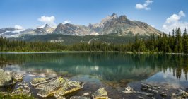 Vue panoramique de la chaîne de montagnes qui se reflète sur un lac alpin avec un rivage rocheux et un ciel et des nuages bleus ; Colombie-Britannique, Canada — Photo de stock
