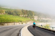 Female cyclist along river pathway with rolling hillside vineyards and mist in the river valley, North of Remich; Luxembourg — Stock Photo