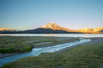 Ein Bach, der zu einem See in großer Höhe führt. Ein Vulkan und schneebedeckte Berge in der Ferne werden vom Sonnenaufgang vor einem reinblauen Himmel erleuchtet; Mendoza, Argentinien — Stockfoto