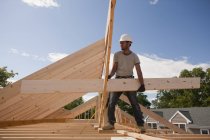 Carpintero que lleva una viga de techo en el sitio de construcción del edificio - foto de stock