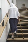 Vista trasera de una monja subiendo por una escalera con la ayuda de un bastón - foto de stock