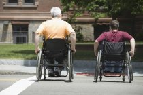 Вид сзади на зрелого мужчину с мышечной дистрофией и молодую женщину, переходящую дорогу в инвалидных колясках — стоковое фото