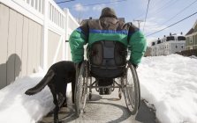 Mujer con esclerosis múltiple en silla de ruedas con un perro de servicio subiendo por la calle nevada - foto de stock