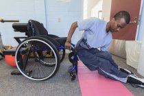 Hombre que tenía meningitis espinal subiéndose a su silla de ruedas desde una almohadilla de yoga - foto de stock