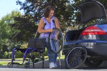 Женщина со Спиной Бифидой с помощью костылей разбирает инвалидное кресло на части для передвижения в машине — стоковое фото