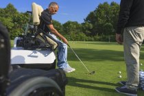 Человек с травмой спинного мозга в адаптивной тележке в гольф положить зеленый с инструктором — стоковое фото