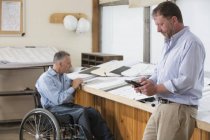 Два инженера проекта используют свои таблетки для проверки планов работы, один в инвалидной коляске с травмой спинного мозга — стоковое фото