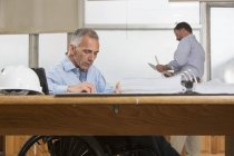 Два инженера-проектировщика оформляют документы о работе, один в инвалидном кресле с травмой спинного мозга — стоковое фото