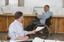 Dos ingenieros de proyecto haciendo papeleo sobre el trabajo, uno en silla de ruedas con una lesión en la médula espinal - foto de stock