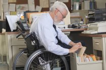 Uomo con distrofia muscolare su una sedia a rotelle cercando scartoffie nel cassetto del suo ufficio — Foto stock