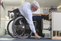 Человек с мышечной дистрофией в инвалидном кресле тянется за чем-то, что он уронил на рабочем столе — стоковое фото