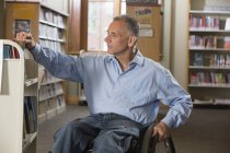 Mann im Rollstuhl mit Querschnittslähmung sortiert Bücher in einer Bibliothek — Stockfoto