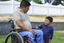 Hombre hispano con lesión de médula espinal en silla de ruedas con su hijo en el césped - foto de stock
