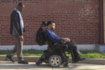 Hombre afroamericano con parálisis cerebral usando su silla de ruedas eléctrica afuera con su asistente de cuidado personal - foto de stock